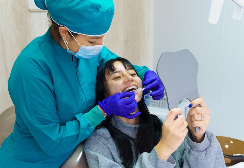 Orthodontist fits braces on smiling patient holding mirror at clinic in Southfield, MI.